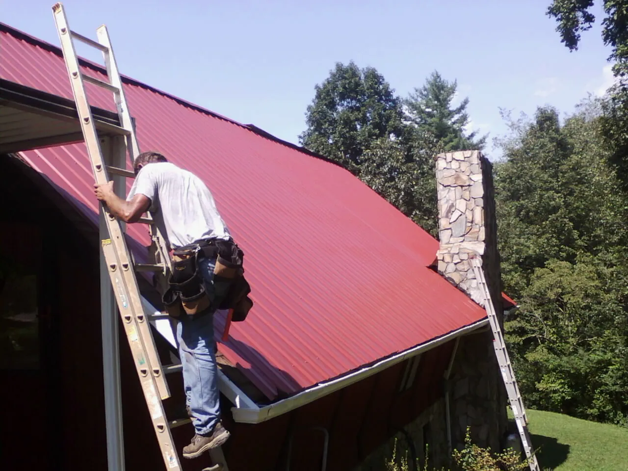 Roofer on a ladder beside a red metal roof and stone chimney — Soffit & Fascia Repair by Roofing Brothers in Schiller Park